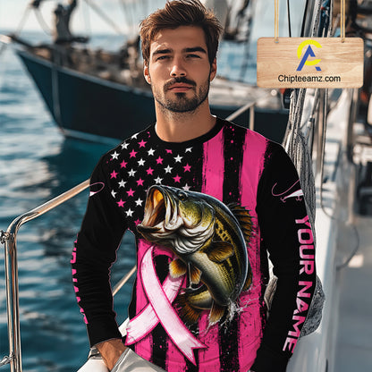 A man wearing a long-sleeve custom shirt with a pink and black striped design, a black American flag pattern on the upper chest, a large bass graphic over a pink ribbon, and 'YOUR NAME' printed down one sleeve. The background shows a blurred marina setting with boats and water.