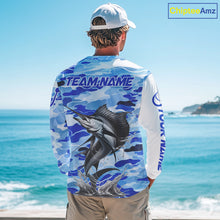 Load image into Gallery viewer, A man wearing a sailfish fishing shirt long sleeve with a large fish graphic and 'TEAM NAME' text, standing by the ocean.